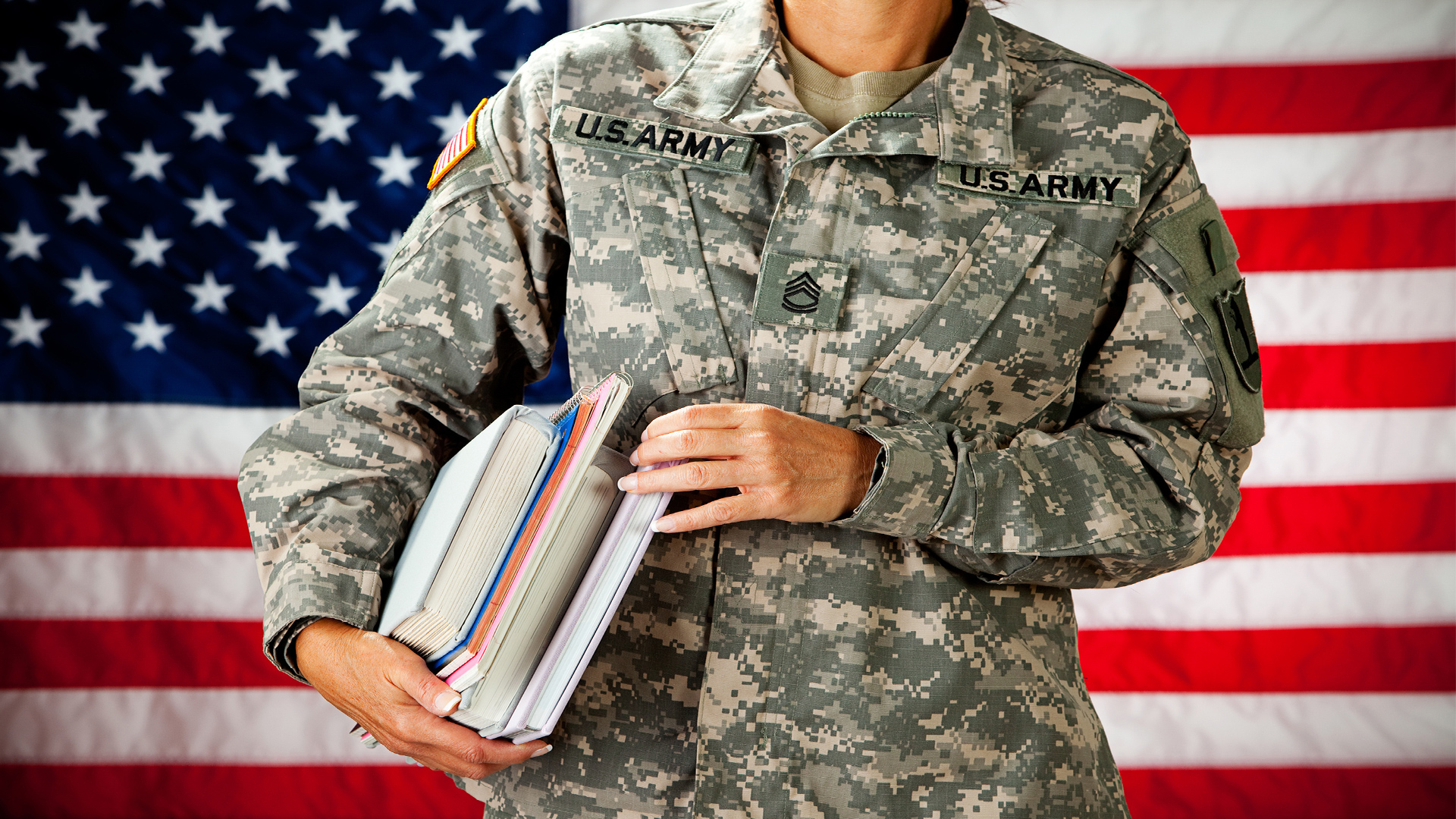soldier in fatigues holding books in front of american flag background
