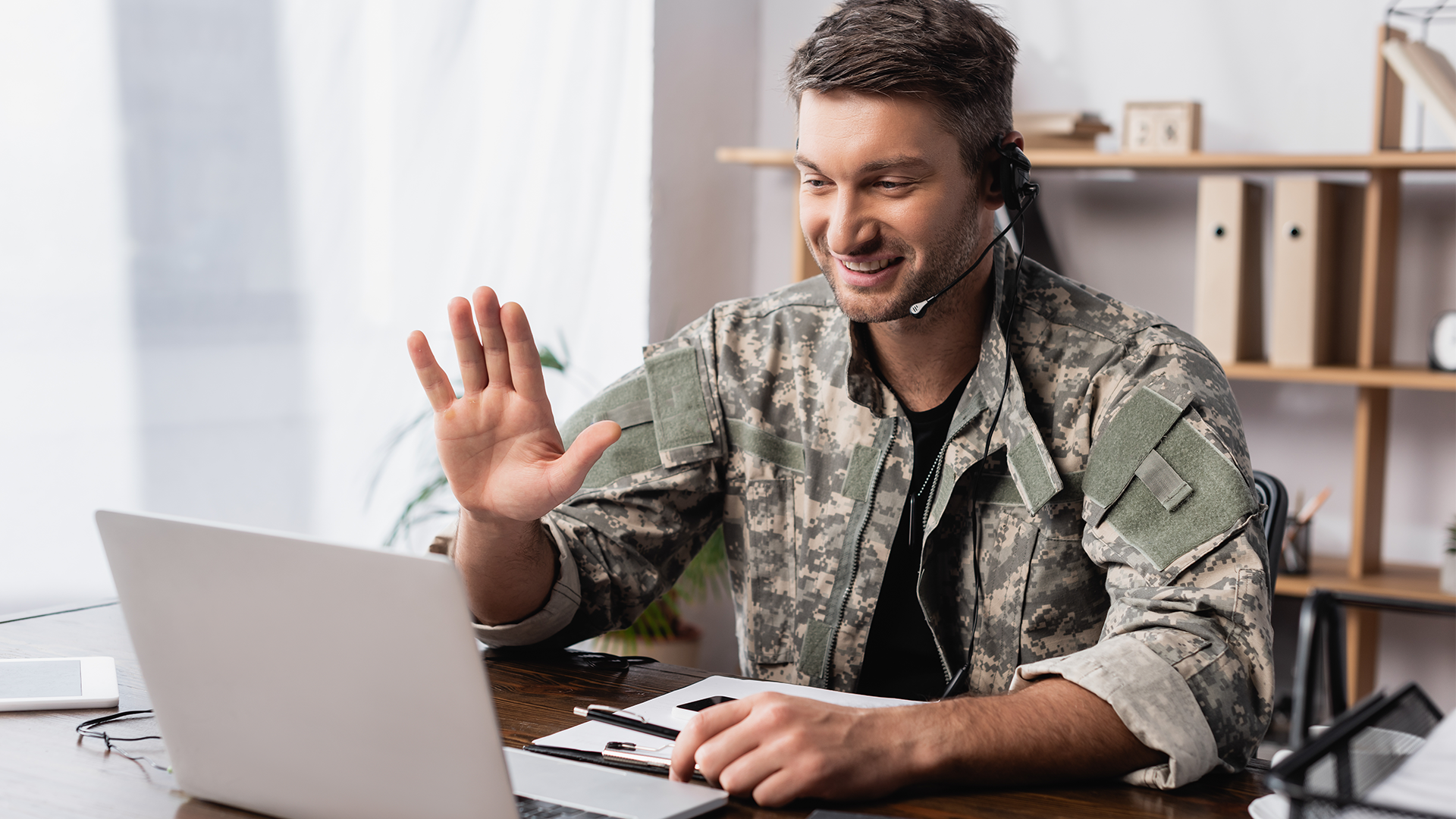 man in fatigues using laptop with headset and waving
